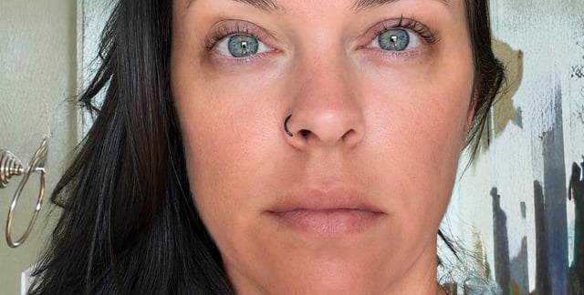 Close-up portrait of a woman with long black hair and blue eyes looking directly at the camera against a white wall background