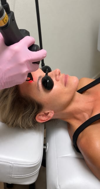 Person receiving laser eye treatment wearing protective goggles while wearing pink gloves applies device near patients face