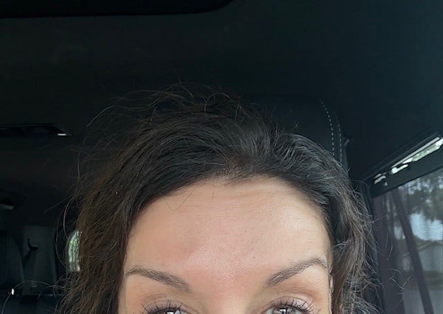 Close-up portrait of a womans face showing her forehead, eyes, and dark hair against a blurred indoor background